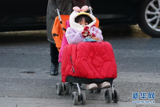 12月29日，在北京朝阳区青年路和朝阳北路路口，一名小朋友坐在盖着厚厚衣服的童车上。当日，入冬最强寒潮来袭，多地迎来今冬最寒冷天气。新华社记者鞠焕宗 摄